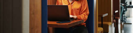 Header of anonymous woman working on laptop in modern office booth, sitting at wooden desk with smartphone nearby, focusing on screen, partial body visible, business environmentの写真素材