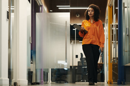 Young Black woman walking through modern office hallway holding digital tablet, looking ahead with focused expression, business environment visible in backgroundの写真素材