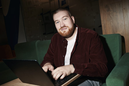 Portrait of Caucasian young adult man with beard looking into camera while sitting on green sofa...
