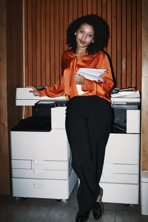 Young Black woman standing beside office printer holding documents, leaning casually against...