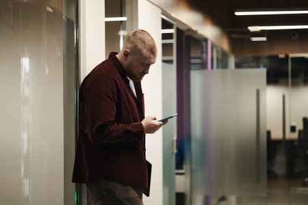 Caucasian young man standing in modern office hallway using smartphone, looking down at screen with...