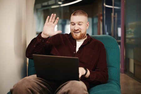 Caucasian young man sitting on chair waving hand while video chatting on laptop smiling and looking...
