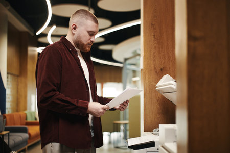 Caucasian man standing at printer holding documents, reviewing printed papers in modern office environment, focused on task, short hair and beard visibleの写真素材