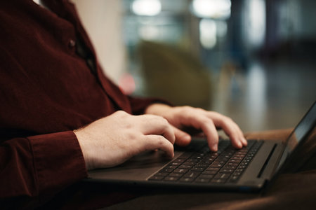 Young man typing on laptop computer with both hands in modern workspace, focusing on keyboard, partial body visible, demonstrating technology use in professional settingの写真素材