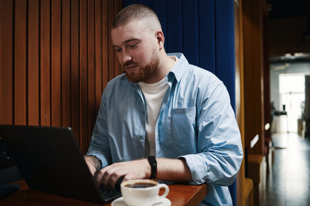 Caucasian young man working on laptop at table in cafe, focusing on screen with hands typing keyboard, short hair and beard visible, coffee cup placed in foregroundの写真素材