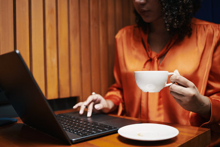 Young woman working on laptop while holding coffee cup, sitting at wooden table with empty saucer, focusing on screen, partial face visible, curly hair framing faceの写真素材