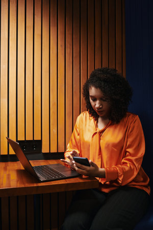 Young Black woman sitting at desk using smartphone while working on laptop, focused expression, curly hair, modern workspace, multitasking with digital devicesの写真素材