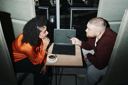 Caucasian young man and Black woman discussing data while sitting at table with laptop displaying charts, both business partners focused on screen, coffee cup on tableの写真素材