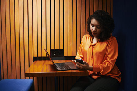 Young Black woman sitting at wooden table using smartphone while working on laptop, looking down at...
