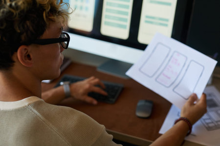 Young adult man sitting at desk working on user interface design, holding paper with smartphone...