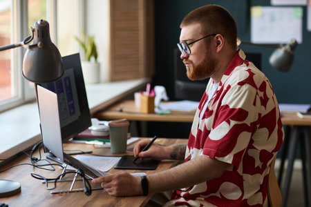 Caucasian young adult man with beard working at desk using desktop computer and digital tablet, focusing on screen while sitting in modern office environment with documents nearbyの写真素材
