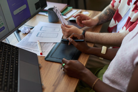 Young adult Black woman and young adult Caucasian man collaborating on digital tablet and reviewing wireframe sketches at desk, working together on user interface design projectの写真素材