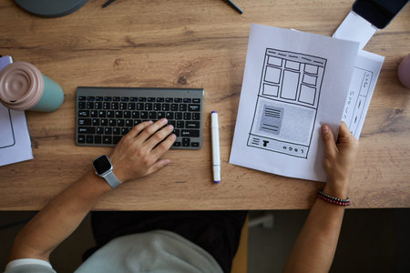 Young adult Caucasian man working at desk holding website wireframe sketch in one hand while typing...