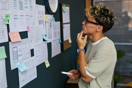 Young adult man analyzing wireframe sketches and user interface designs on wall, holding marker and...