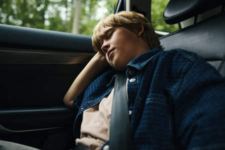 Caucasian boy sleeping in back seat of car during family road trip, head resting against window,...