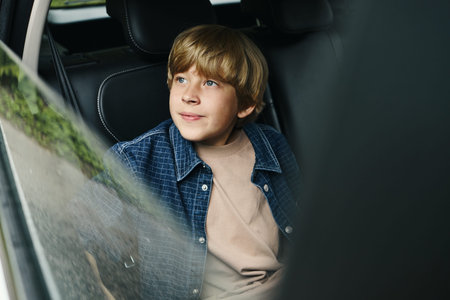 Caucasian boy sitting in car back seat looking out window during family road trip, light brown hair...
