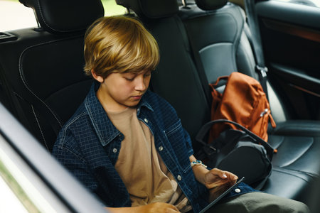 Caucasian teenage boy sitting in back seat of car using digital tablet during family road trip, focused on screen with backpack beside him, natural light coming through windowの写真素材