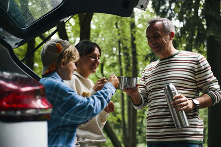 Caucasian middle aged man pouring drink from thermos while smiling with Caucasian woman and Caucasian boy near car trunk during family road trip in forest, all standing outdoorsの写真素材