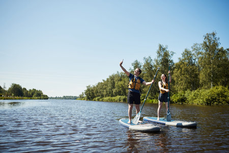 Caucasian young man and Caucasian young woman standing on paddle boards, taking selfie with...