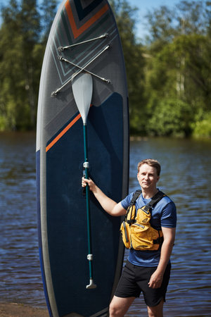 Portrait of Caucasian young adult man standing outdoors holding paddleboard and paddle near river,...