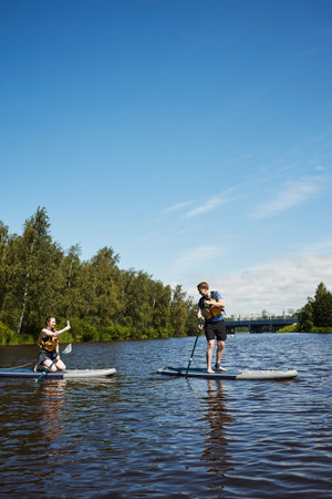 Caucasian young man standing on paddleboard paddling while Caucasian young woman, kneeling on...