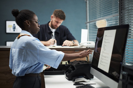 Black female employee working at computer terminal in bank office while Caucasian man standing at...