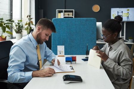 Caucasian male clerk signing documents while Black young woman reviewing paperwork across desk in...