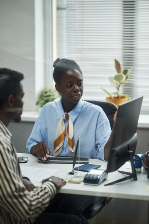 Young female consultant explaining financial information to middle aged Black man in bank office,...