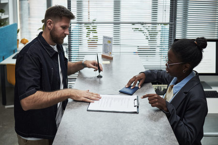 Caucasian young man signing document while Black female banker explaining loan details at counter in...