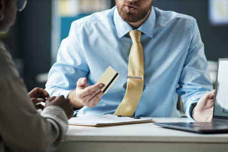 Caucasian male adviser explaining credit card features to Black woman in bank office, both sitting...