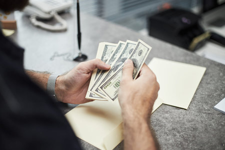 Hands of unrecognizable man counting US dollar bills at bank office desk, holding cash in hands near...