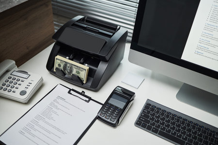 Office desk showing money counting machine processing US dollar bills beside desktop computer,...