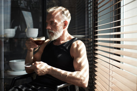 Caucasian middle aged man with gray beard sitting by window holding cup and saucer drinking coffee...