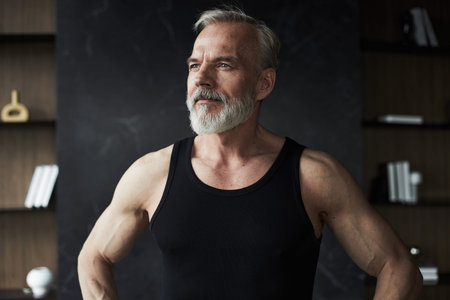 Middle aged man standing with hands on hips, looking away from camera, showing muscular arms and confident posture, short gray hair and full beard visibleの写真素材