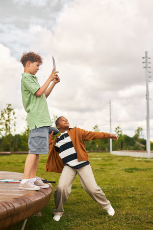 Caucasian boy standing on bench holding digital tablet taking photo while Black boy throwing ball in...