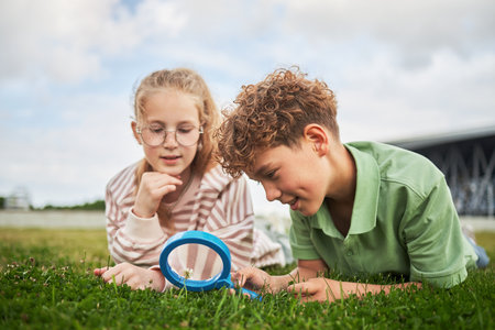 Caucasian girl and Caucasian boy lying on grass examining small object with magnifying glass, girl wearing glasses, boy focusing intently on discoveryの写真素材