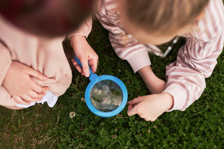 Caucasian girls examining grass and flowers with magnifying glass outdoors, both sitting on green lawn, focusing on nature exploration and discovery togetherの写真素材