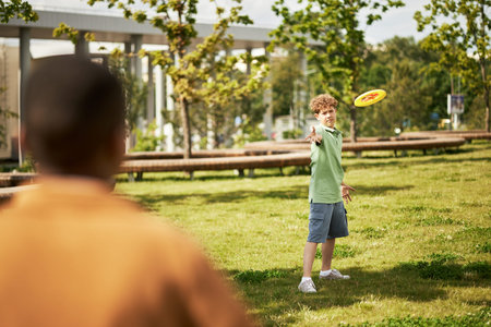 Caucasian boy throwing flying disc toward Black boy in park, both children engaging in outdoor activity on grass with trees and urban structures visible in backgroundの写真素材