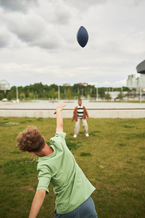 Caucasian child throwing football toward Black friend standing on grass in outdoor park, both...