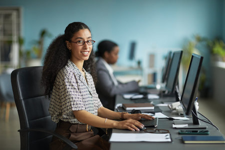 Portrait of young adult woman smiling while working at computer desk in modern office, sitting in...
