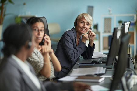Caucasian middle aged woman smiling while wearing headset and sitting at desk with multiethnic young...
