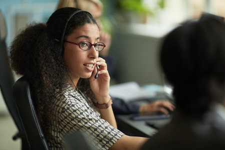 Young adult woman wearing headset talking to colleague in modern office environment, sitting at desk...