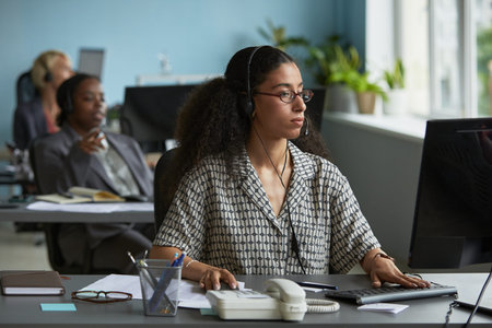 Young adult woman wearing headset working at computer in modern office, sitting at desk with...