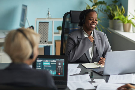 Young adult Black woman wearing headset smiling while participating in online meeting at office...