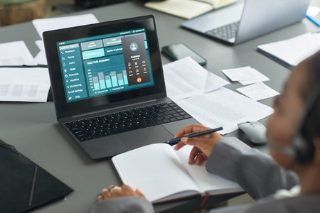 Black woman working at desk analyzing business data on laptop screen while taking notes in notebook,...