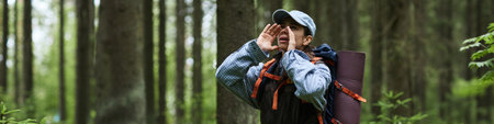 Teenage Caucasian boy standing in forest shouting with hands around mouth wearing backpack and cap, engaging in outdoor activity surrounded by tall trees and greeneryの写真素材