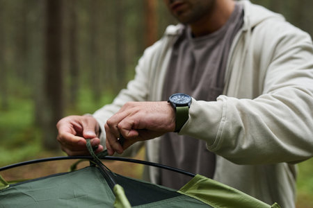 Middle aged Caucasian man assembling tent outdoors in forest, hands tying tent poles, wearing...