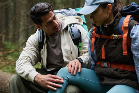 Caucasian young adult man comforting Caucasian young adult woman with injured knee during hiking in forest, both wearing backpacks and outdoor gear, sitting on fallen treeの写真素材