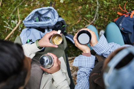 Two young adult multiethnic hikers sitting outdoors holding metal mugs with hot drinks, relaxing together in forest setting with backpacks visible on ground nearbyの写真素材