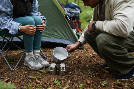 Caucasian young adult man pouring water from metal pot into camping mugs while sitting with...